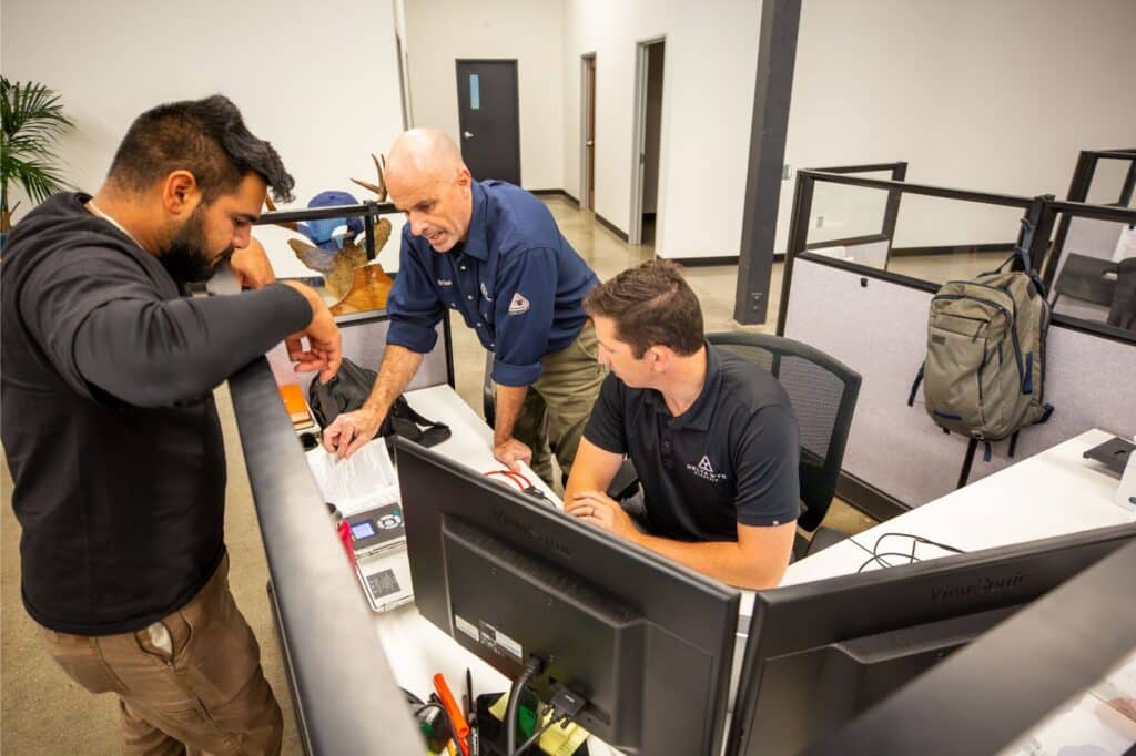 Three men are at a workspace. One stands at a desk talking to a seated man who, perhaps collaborating with an industrial electrical contractor, is working on a computer. Another stands and points at a document on the desk. Office cubicles and a backpack are in the background.